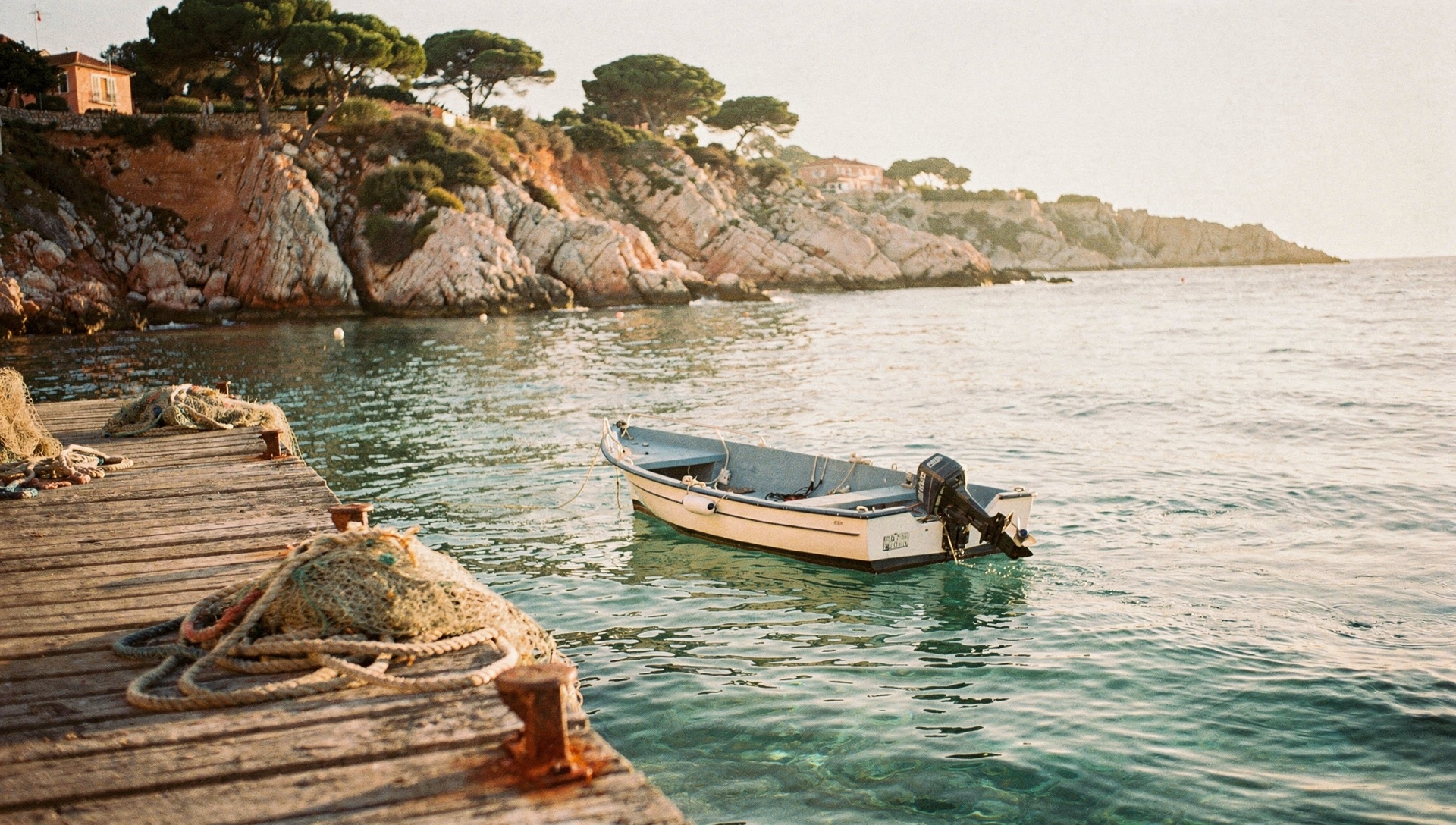 Barca de alquiler amarrada junto a un muelle de madera del puerto de Blanes, con una cala de roca y pinos de la Costa Brava al fondo al atardecer