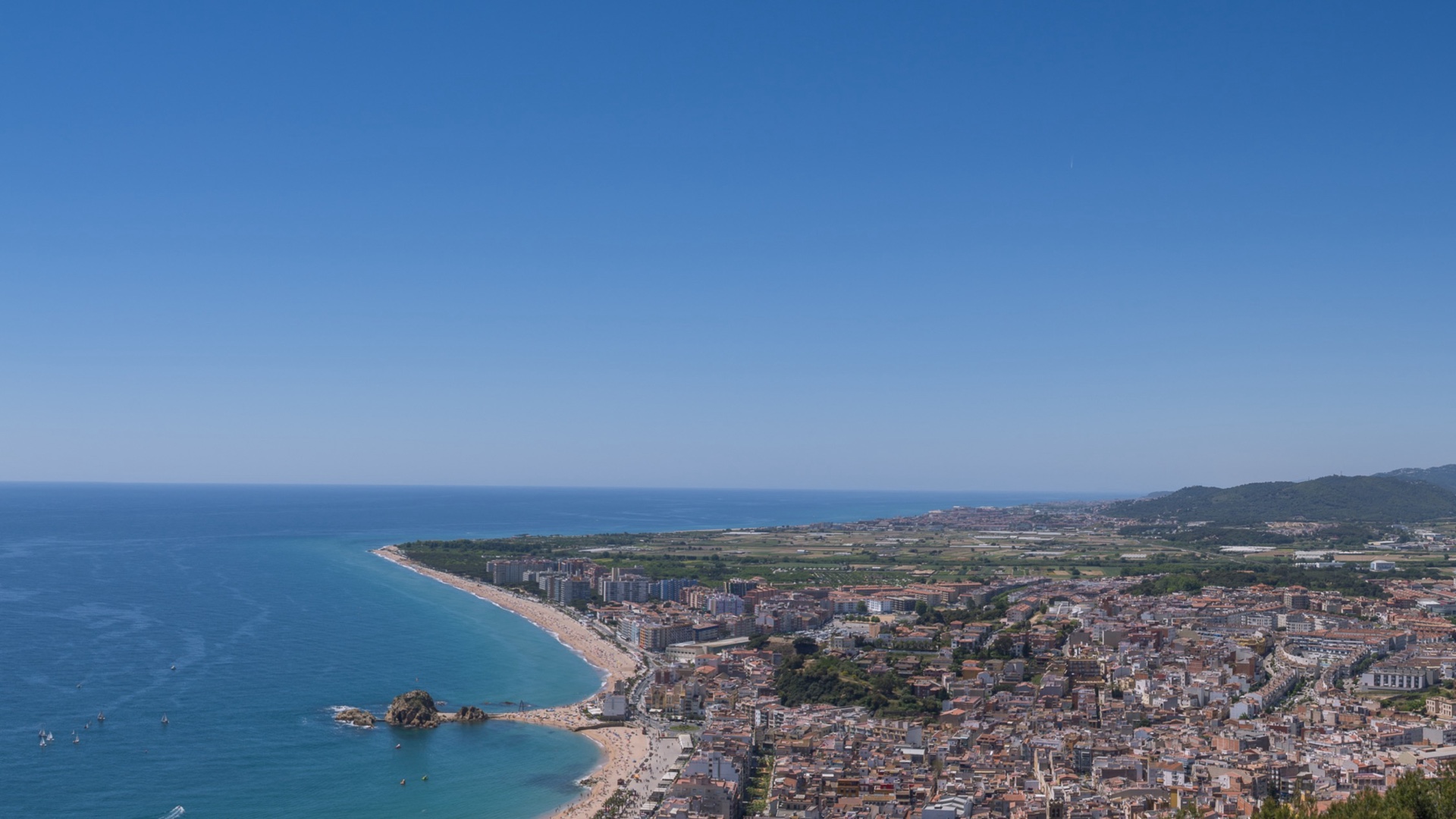 Vue panoramique depuis le Castell de Sant Joan de Blanes montrant Sa Palomera, la longue plage de S'Abanell, le centre-ville et les plaines du Maresme au loin sous un ciel bleu