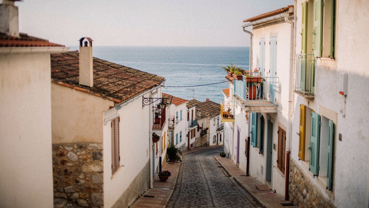Calle estrecha del casco antiguo de Blanes con casas mediterráneas bajas, tejas de terracota, contraventanas de madera en colores pastel y plantas en pequeños balcones a la luz dorada de la tarde