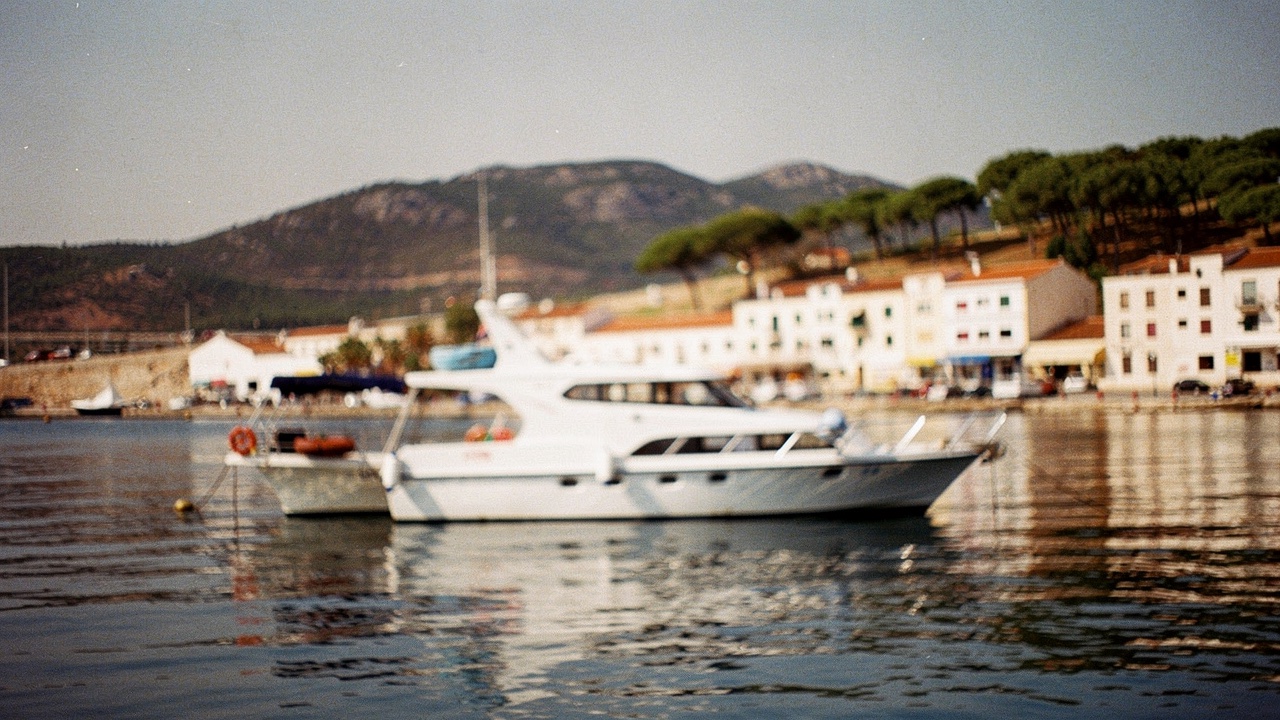 Grand bateau type catamaran touristique amarré dans un petit port méditerranéen avec maisons blanches et collines de pins en arrière-plan à la lumière chaude du matin