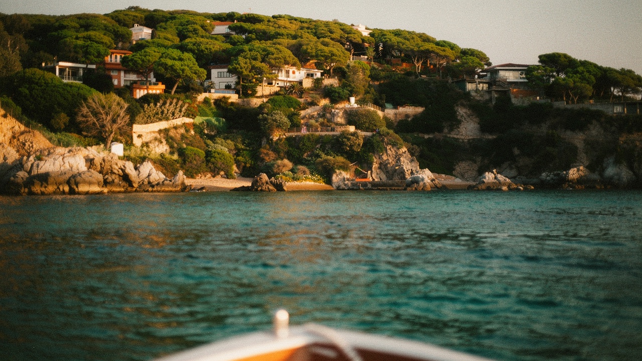 Vista desde un barco navegando junto a la costa de Blanes, con una cala escondida entre acantilados de roca, pinos mediterráneos y aguas turquesa al atardecer