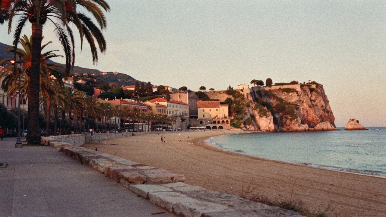 View of the Blanes seafront with a long curving golden-sand beach, palm trees, low buildings and a rocky outcrop visible in the middle of the bay in warm afternoon light