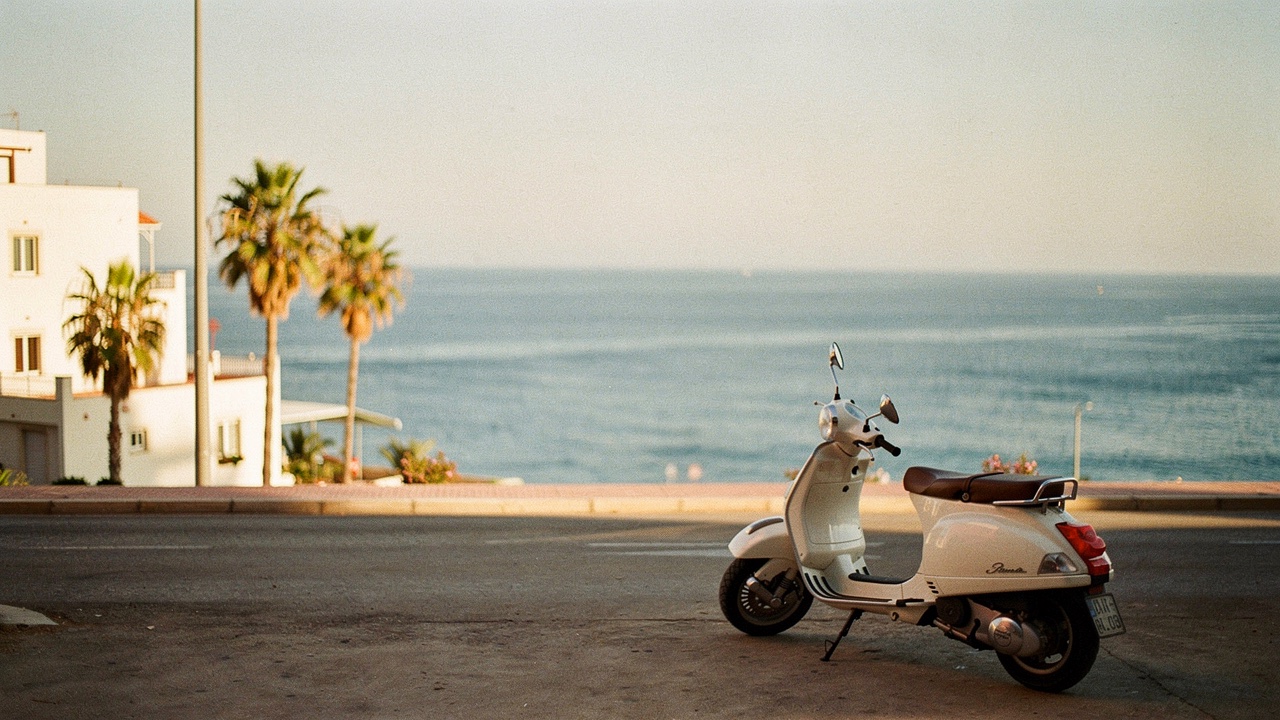Scooter blanc garé dans une rue côtière méditerranéenne avec la mer en fond, maisons blanches basses et palmiers à la lumière dorée du coucher du soleil