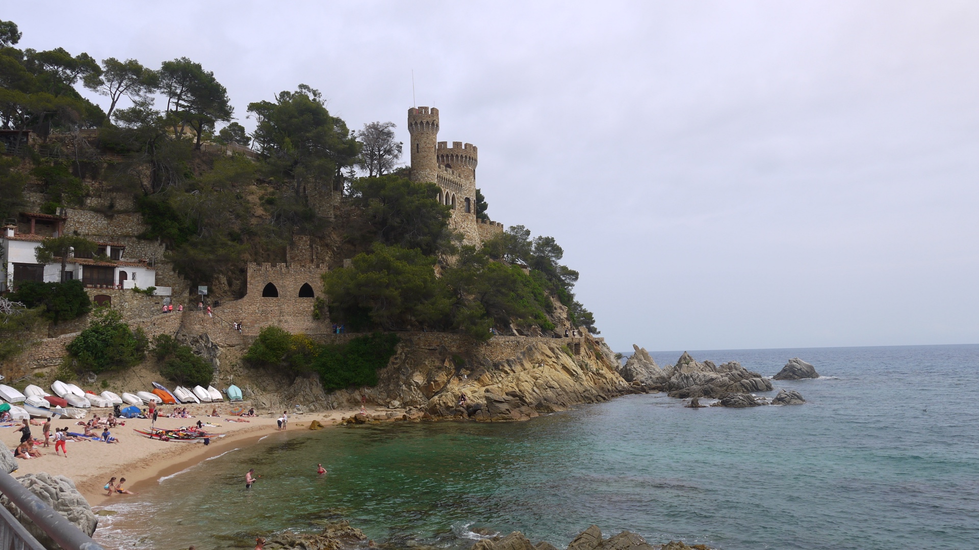 Castell d'en Plaja à Lloret de Mar vu depuis la plage de Sa Caleta, avec ses tours crénelées sur une falaise rocheuse, pins entourant la base et la crique avec barques au premier plan