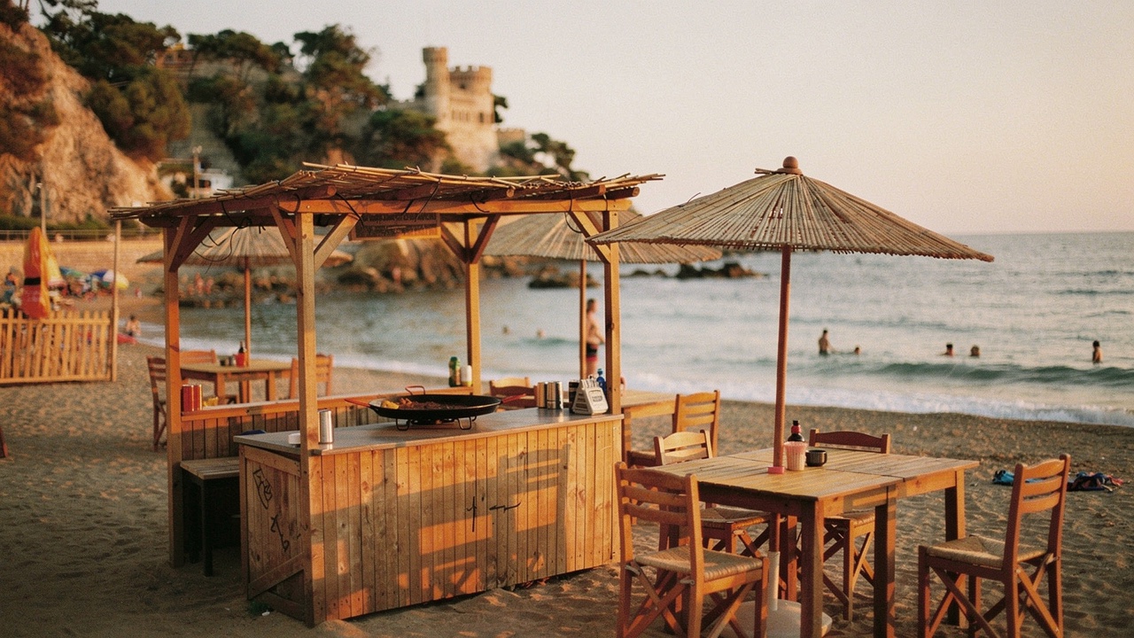Chiringuito tradicional mediterráneo en la arena al atardecer con mesas de madera bajo sombrillas de bambú, una paella sobre la mesa y mar tranquilo al fondo a la luz dorada