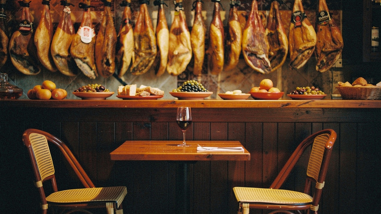 Intimate tapas bar interior in Lloret de Mar with Iberian hams hanging above a wooden counter, small plates of olives and bread, a glass of red wine and wicker chairs