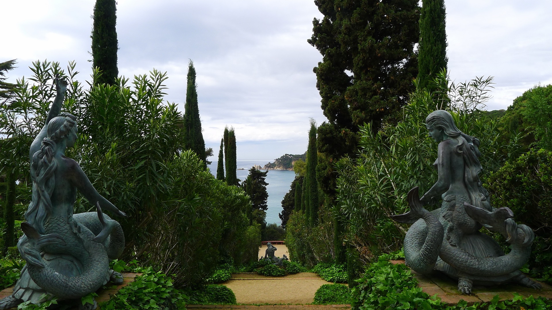 Vista de las esculturas de las Sirenas de los Jardines de Santa Clotilde en Lloret de Mar, con cipreses italianos enmarcando el mar Mediterráneo al fondo en un día gris suave